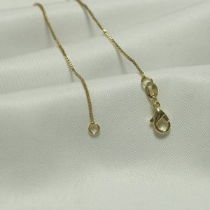May include: A delicate gold-colored necklace chain with a lobster clasp and a small ring at the end. The chain is displayed on a white, textured fabric, highlighting its fine details and elegant design.