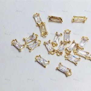 May include: A collection of gold-toned metal charms featuring clear rectangular stones. Each charm has a loop on either end for attaching to jewelry or other crafts.