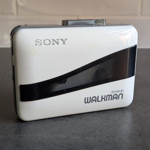 Può includere: Un lettore di cassette Sony Walkman bianco con una striscia diagonale nera. La parola "SONY" è stampata in alto e "WALKMAN" in basso in lettere grigie. Il lettore ha un pulsante argentato.