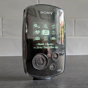 May include: A black Sony Walkman digital media player with a glossy finish. The screen displays a menu with icons and the text Music Library and ez The Killer. Below the screen is a circular navigation pad, an Option button, and a Back button.