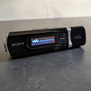 May include: A black Sony Walkman digital music player with a rectangular body, a small digital screen displaying the Walkman logo in blue, a circular play-pause button, and a small back button. The device features silver accents and branding.