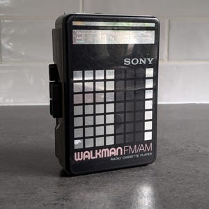 Può includere: Lettore di cassette radio FM/AM Sony Walkman nero. Il dispositivo presenta un design a griglia argentata e nera sul davanti, con la scritta "WALKMAN" e "FM/AM RADIO CASSETTE PLAYER" stampata in bianco. Il dispositivo è rettangolare e ha una clip sul lato.