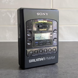 Op de afbeelding: Een zwarte Sony Walkman FM/AM draagbare cassettespeler met een digitaal display, Mega Bass-instellingen en auto-reverse functionaliteit. Het apparaat bevat diverse bedieningsknoppen, een geheugenfunctie en een bandselector voor radio-ontvangst.