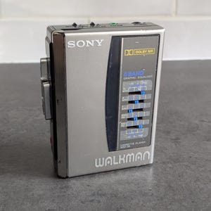 Könnte beinhalten: Ein silberner Sony Walkman WM-30 Kassettenrekorder mit schwarzer Blende, 5-Band-Grafik-Equalizer, Dolby NR-Logo und blauen Schiebereglern. Das Gerät hat eine metallische Oberfläche mit deutlich sichtbaren Beschriftungen und seitlichen Tasten.