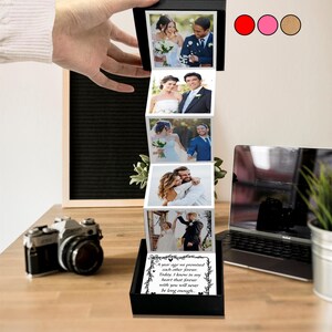 May include: A black photo box displaying a series of wedding photographs, unfolding to reveal images of a couple. The box includes a printed message and is on a wooden surface. A vintage camera and laptop are also visible.