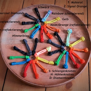 May include: A variety of colorful paracord knots, including black and charcoal diamonds, neon green, and rainbow, displayed on a wooden surface. The knots are labeled with their names in German, including "Schlangenknoten" (snake knot), "H&ouml;hlenknoten" (cave knot), and "Diamantknoten" (diamond knot).