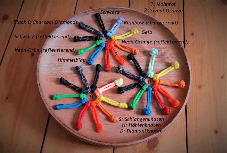 May include: A variety of colorful paracord knots, including black and charcoal diamonds, neon green, and rainbow, displayed on a wooden surface. The knots are labeled with their names in German, including "Schlangenknoten" (snake knot), "H&ouml;hlenknoten" (cave knot), and "Diamantknoten" (diamond knot).