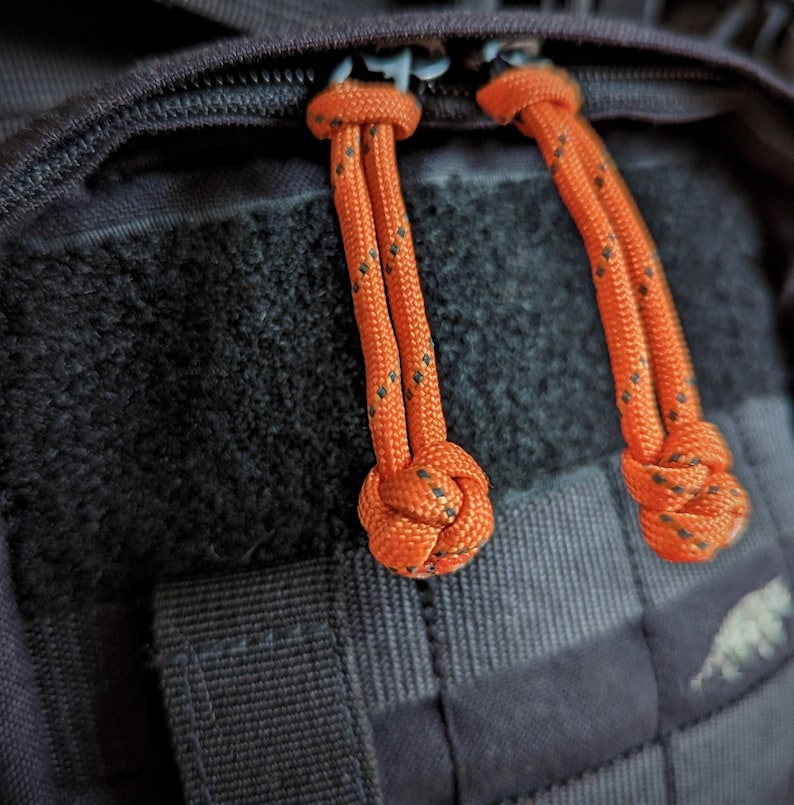 May include: Two orange zipper pulls with black accents are attached to a black zipper on a dark gray backpack.