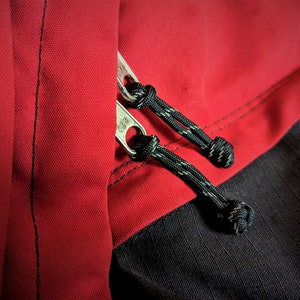 May include: Close-up of a red and black jacket with a zipper and black drawstrings. The zipper has a silver pull tab.