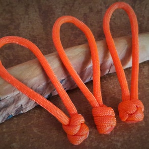 May include: Three orange paracord keychains with a knot at the end of each. The keychains are on a piece of wood.