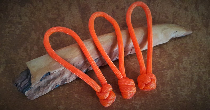 May include: Three orange paracord keychains with a knot at the end of each. The keychains are on a piece of wood.