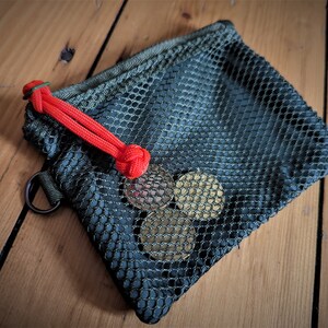 May include: A small green mesh drawstring bag with a red cord closure. The bag is filled with four gold-colored coins.