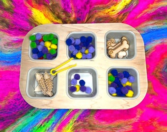 May include: A wooden tray with six compartments, each containing different items. The compartments contain purple, blue, and yellow pom-poms, two wooden fish, two wooden bones, and a yellow plastic tool.