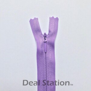 K&ouml;nnte beinhalten: Ein lavendelfarbener Rei&szlig;verschluss mit geschlossenem Schieber. Die Rei&szlig;verschlussz&auml;hne sind oben sichtbar, und der Stoff hat ein Fischgr&auml;tenmuster. Der Text "Deal Station" ist unten aufgedruckt.
