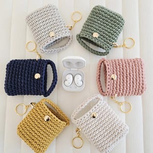 May include: A collection of handmade crochet earbud cases in various colors, including gray, green, navy, pink, yellow, and cream. Each case has a gold-colored button closure and a matching keychain ring. The earbuds are in a white case.