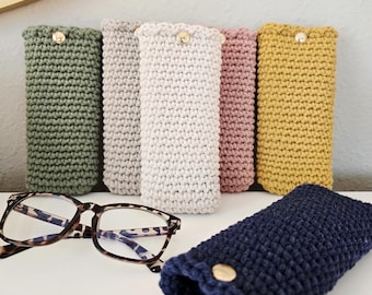 May include: A collection of colorful, crocheted eyeglass cases in shades of green, gray, cream, pink, and yellow, each with a gold button closure. A pair of eyeglasses with a tortoiseshell frame rests beside the cases, and a navy blue case is in the foreground.