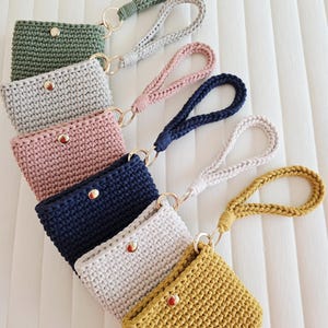 May include: A collection of handmade crochet wristlet wallets in various colors, including green, gray, pink, navy, white, and yellow. Each wallet features a button closure and a matching wrist strap with a gold-colored ring.