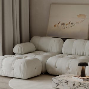 May include: A modern living room with a large, off-white tufted sofa and ottoman. A framed artwork depicts animals walking towards a boat. A marble-topped coffee table holds a lamp and a book. Soft, neutral tones create a calming atmosphere.
