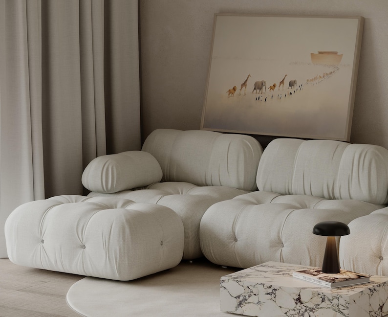 May include: A modern living room with a large, off-white tufted sofa and ottoman. A framed artwork depicts animals walking towards a boat. A marble-topped coffee table holds a lamp and a book. Soft, neutral tones create a calming atmosphere.