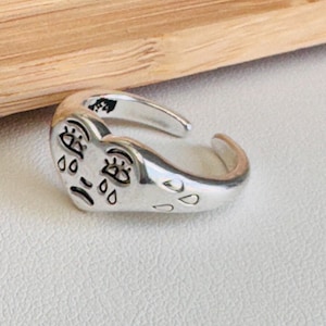 Puede incluir: Anillo de plata con un diseño en forma de corazón que presenta una cara llorando con detalles de lágrimas. El anillo es ajustable.