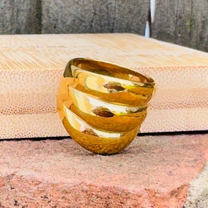 May include: A gold ring with a distinctive design. The ring has a series of horizontal, curved bands that encircle the finger. The bands have a reflective surface, creating highlights and shadows. The ring is set against a neutral background.