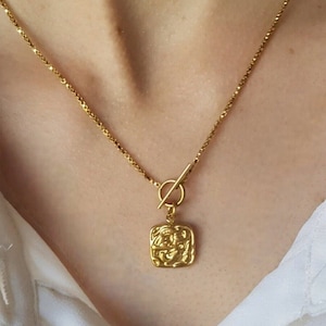 Op de afbeelding: Een gouden ketting met een vierkante hanger met een ingewikkeld ontwerp. De ketting heeft een toggle-sluiting.