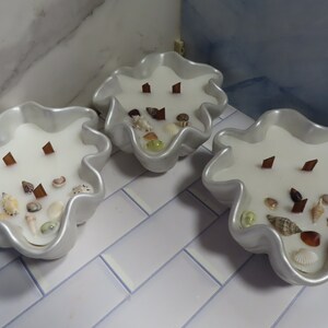 May include: Three white seashell-shaped candles with wooden wicks and embedded seashells and decorative stones. The candles are in pearlescent containers and sit on a white and blue tiled surface. The candles are a decorative home accent.