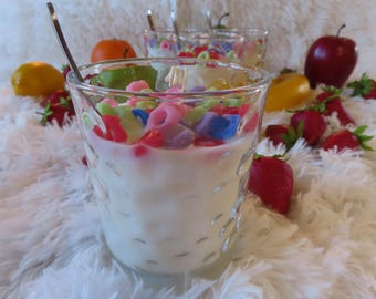 May include: A glass filled with a white candle that looks like milk and colorful cereal. The candle is surrounded by fruit and other decorative items.