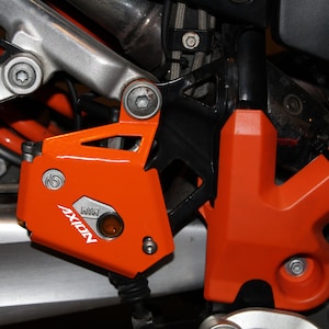 May include: Close-up of a motorcycle engine guard with an orange AXION logo and a black and orange plastic cover. The guard is made of metal and has a silver finish.