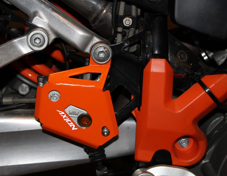 May include: Close-up of a motorcycle engine guard with an orange AXION logo and a black and orange plastic cover. The guard is made of metal and has a silver finish.