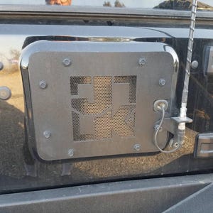 Puede incluir: Panel metálico negro con un diseño perforado y las letras "FK" en la parte trasera de un vehículo. El panel está asegurado con pernos y tiene un soporte para antena.