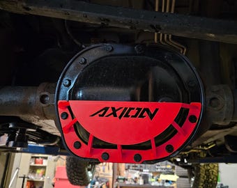May include: A red metal differential cover with the word "AXION" printed in black. The cover is attached to a black metal differential housing.