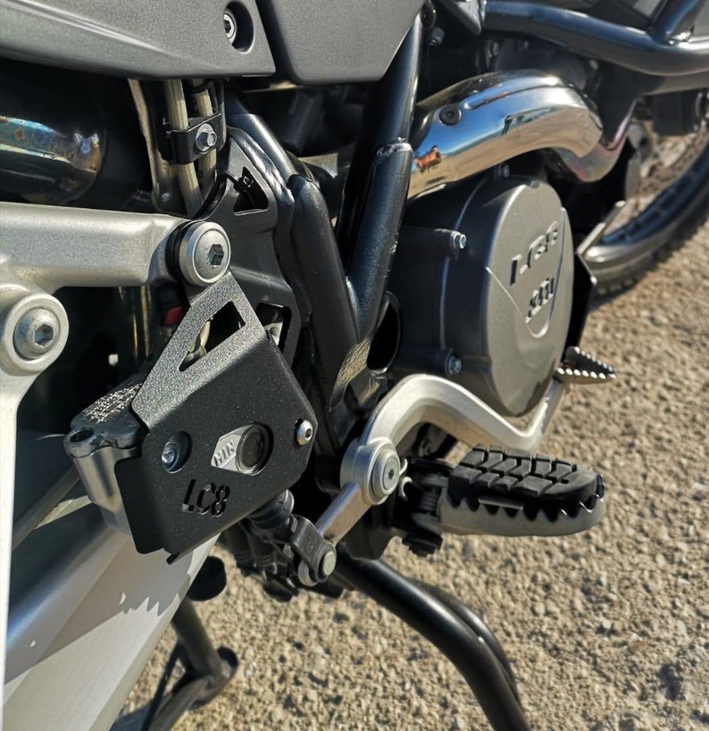 May include: Close-up of a black motorcycle engine guard with the text "LC8" on it. The guard is attached to a silver motorcycle frame.