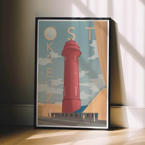 Może przedstawiać: Oprawiony wydruk artystyczny z ilustracją czerwonej latarni morskiej i złotym napisem "OSTKREUZ". Grafika ma styl vintage plakatu podróżniczego, na tle jasnoniebieskiego nieba z chmurami. Czarna rama.