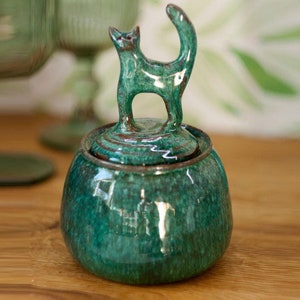 Peut inclure: Un pot en céramique vert avec un couvercle orné d'une figurine de chat. Le pot a un glaçage moucheté et une finition brillante.