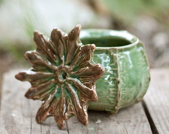 May include: A handmade ceramic jar with a lid. The jar is a muted green color with a textured surface. The lid is a flower shape with brown and green accents. The jar is sitting on a wooden surface.