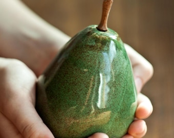 May include: A ceramic pear with a green glaze and a brown stem. The pear is sitting in the hands of a person.