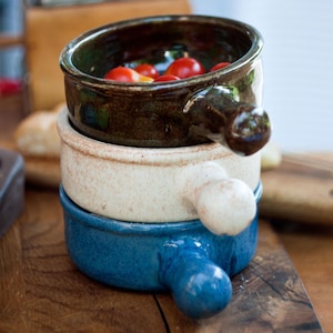 May include: Three ceramic bowls stacked on top of each other, each with a handle. The top bowl is dark brown and filled with red cherry tomatoes. The other bowls are off-white and blue. The bowls are on a wooden surface.