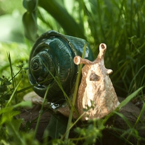 Könnte beinhalten: Eine Keramik-Schnecken-Skulptur mit einer grünen Schale und einem braunen Körper. Die Schnecke sitzt auf einem Bett aus grünem Gras mit roten Tulpen im Hintergrund.