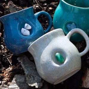 Könnte beinhalten: Drei Keramikbecher mit einzigartigen Designs. Zwei Becher sind blau mit weißen Steinen, die in die Seite eingebettet sind. Ein Becher ist weiß mit einem grünen Stein, der in die Seite eingebettet ist.