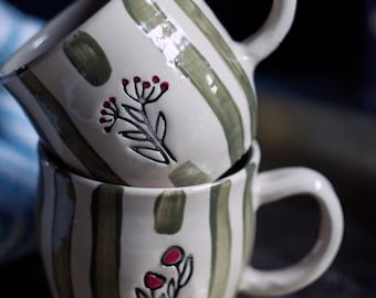 Puede incluir: Dos tazas de cerámica apiladas con patrones de rayas blancas y verdes. Cada taza presenta un diseño floral pintado a mano con detalles en rojo. Las tazas tienen asas grandes y redondeadas.