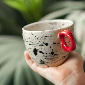 Könnte beinhalten: Eine Keramik-Tasse mit weißem Grund und schwarzem Spritzmuster. Die Tasse hat einen leuchtend roten Griff. Die Tasse wird in einer Hand gehalten, mit einem verschwommenen grünen Hintergrund.