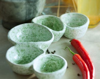 May include: A set of four small, speckled green and white ceramic serving bowls. Two vibrant red chili peppers and a mortar and pestle are also visible. The bowls are arranged on a light-colored surface, suggesting a culinary theme.