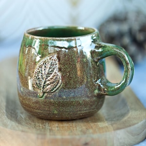 May include: A green ceramic mug with a brown glaze and a leaf design. The mug has a rounded body and a handle.