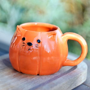 Może przedstawiać: Pomarańczowy ceramiczny kubek w kształcie dyni z kocimi cechami. Kubek ma uchwyt i małe uszy. Czarne kropki tworzą oczy i nos, z wąsami. Kubek stoi na drewnianej powierzchni.