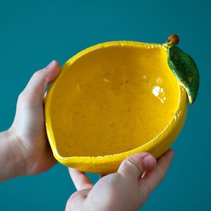 Könnte beinhalten: Eine gelbe Keramikschüssel in Form einer Zitrone mit einem grünen Blatt oben. Die Schüssel wird von zwei Händen gehalten.