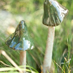 Puede incluir: Dos esculturas de hongos de cerámica con sombreros cónicos de color verde oscuro brillante con crestas verticales visibles y tallos delgados, texturizados y de color blanco hueso. Los sombreros tienen un acabado esmaltado reflectante.