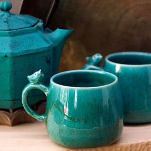 Può includere: Due tazze in ceramica blu turchese con manici a forma di uccello. Le tazze sono su una superficie di legno.