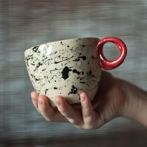Może przedstawiać: Ceramiczny kubek z kremową podstawą i czarnym wzorem rozprysków farby. Kubek ma jaskrawoczerwony uchwyt. Kubek jest trzymany w dłoni osoby.
