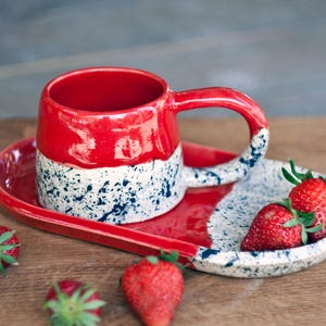 Può includere: Un set di tazza e piattino in ceramica fatto a mano. La tazza e il piattino sono rossi con un motivo maculato bianco e blu. Fragole fresche sono poste sul piattino. La tazza ha un manico grande.